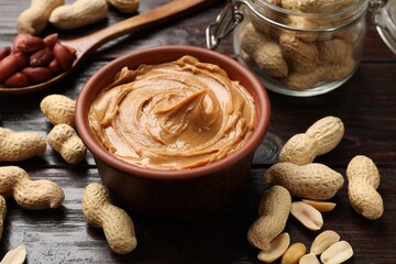 Tasty peanut butter in bowl, groundnuts, jar and spoon on wooden table, closeup