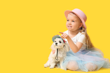 Cute little girl pointing at something with Maltese dog in cap and sitting on yellow background