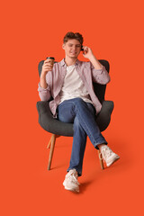 Handsome young man with paper cup of coffee talking by mobile phone and sitting in armchair on orange background