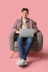 Handsome young man with modern laptop sitting in armchair on pink background