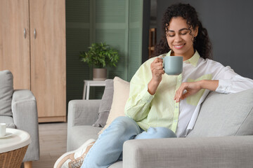 Young African-American woman with cup of tea resting on sofa at home