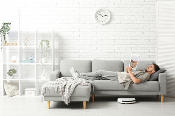 Modern robot vacuum cleaner near sofa with resting young man at home