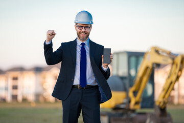 Civil engineer worker at a construction site. Mature engineer worker. Man in suit and hardhat helmet at construction site. Middle aged head civil engineer worker standing outside near excavator.