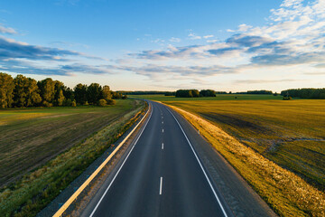 A winding road stretches into the distance, flanked by vibrant green fields and dense trees under a clear blue sky.