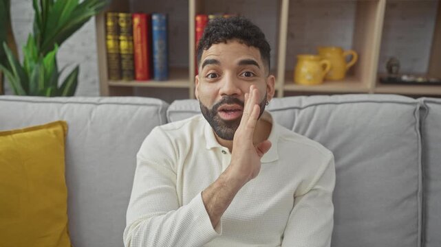 Young hispanic man whispering a malicious rumor, secret talk and gossip conversation at home, hand covering mouth