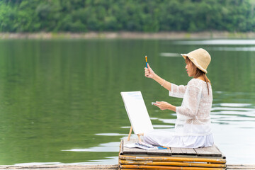 Young Asian woman enjoy and fun outdoor lifestyle travel nature forest mountain on summer holiday vacation. Happy girl relaxing with hobbies drawing and painting watercolor on lake house balcony.