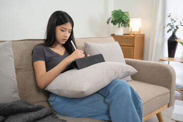 Obraz premium Young asian women reading business report about marketing and finance data of project and taking notes in tablet while sitting on couch to spending time and relaxation with enjoying lifestyle at home
