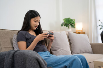 Young asian gamer women using smartphone to playing video game with having fun and happiness while sitting on comfortable couch to spending time and relaxation with enjoying lifestyle at home