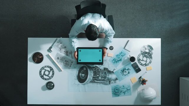 Top view of skilled engineer writing in paper while looking at tablet display green screen with turbine engine sketch design and metal gear and architectural equipment scatter around. Alimentation.