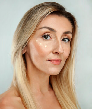Beautiful gorgeous 50s mid middle aged mature woman with three dots of eye cream on face isolated on white background. Mature european lady portrait. Old Age Concept for Skincare, Cosmetics Product