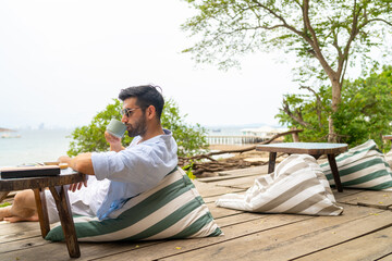 Happy Caucasian man enjoy outdoor lifestyle travel tropical island on summer beach holiday vacation. Handsome guy relaxing and drinking coffee at beach cafe and looking beautiful nature of the sea.