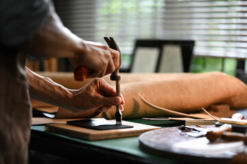 Obraz premium Asian male artisan carefully punching piece of leather at professional leather workshop