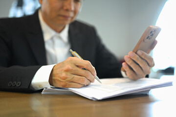 Senior businessman holding mobile phone while examining a financial report at his office desk