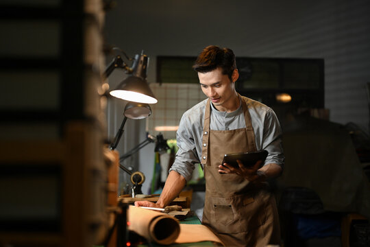 Handsome male leather artisan wearing apron using digital tablet at his workbench surrounded by leather rolls and tools