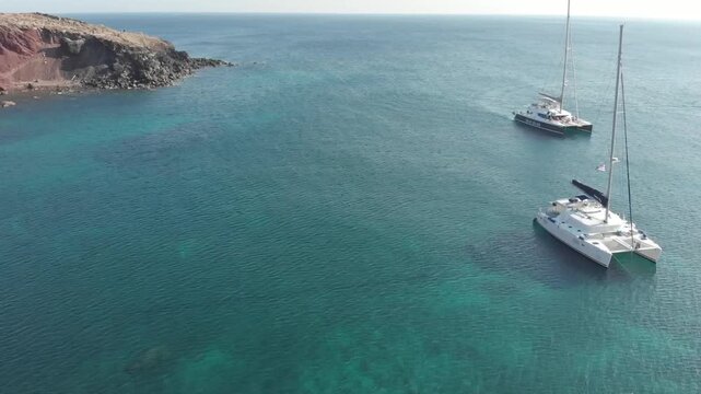 Aerial panorama drone view of white yachts sail in the sea with tourists near Red beach in Santorini in Akrotiri region, Greece. Aegean Sea, Mediterranean. From above

