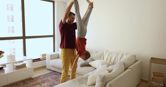 Happy strong Hispanic dad lifting kid upside down, holding hanging boys legs. Positive active loving father playing with little child, entertaining kid, enjoying family weekend at home