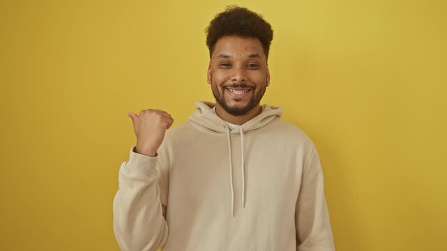 Cheerful african american man wearing a hoodie, flashes a broad smile while pointing to the side with thumb up - looking absolutely chuffed on an isolated yellow background
