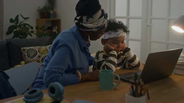 Medium shot of caring mother of African American ethnicity explaining topic of subject to son sitting in front of wireless laptop at home desk