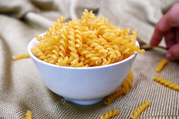 Fusilli pasta in a white bow on wavy fabric background. Close-up fusilli texture in a bow. Concept of raw ingredients, home-cook, pasta, Italian food, and kitchenware. Raw fusilli pasta texture.