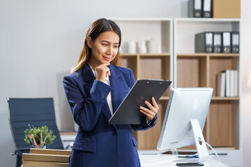 An Asian businesswoman sits at a table, working with a smile. She manages her online Kuya business...