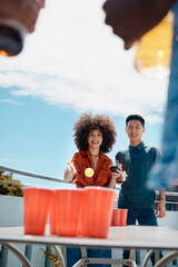 Happy, friends and play beer pong on rooftop to celebrate Octoberfest. Man, woman and throw ball in cup with alcohol on table at party for competition, game or drink wine with diverse people outdoor