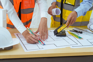 Two young women work at a desk, meticulously designing and drawing plans for building a house. As...