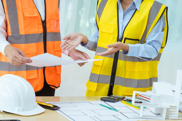 Two young women work at a desk, meticulously designing and drawing plans for building a house. As...