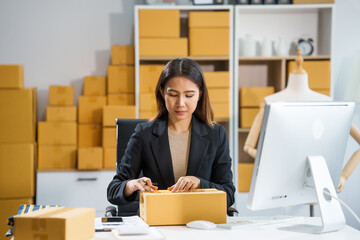 A young Asian businesswoman sits at a table, working on selling products online. She takes orders, packs items, and prepares shipments for customers, ready to send them to the post office.