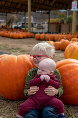 Boy with red glasses holds baby and sits in front of pumpkins