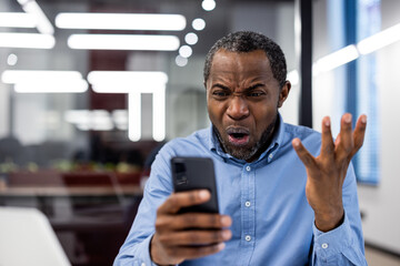 Office worker in blue shirt displaying frustration and disbelief while looking at smartphone in modern office setting. Man showing strong emotion, concept of technology issues or bad news.