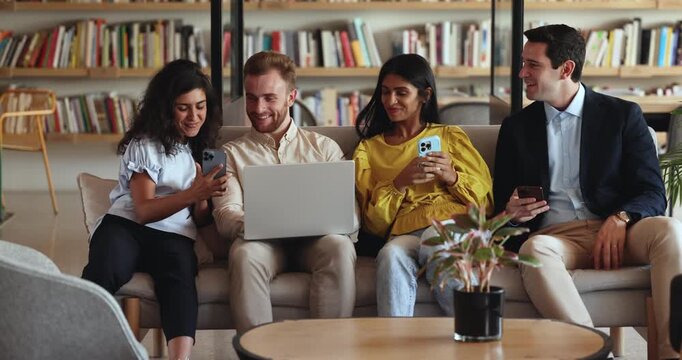 Cheerful diverse team of friends using gadgets in co-working space, showing online content to each others, discussing social media news, funny videos. Group of happy students enjoying break in library
