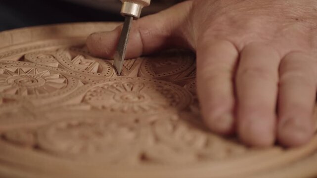 The hand of a man wood carving master holds a cutting tool to applying floral patterns and ornaments on a wooden circle. The concept of handicraft as a hobby, skill, talent and exquisite artwork
