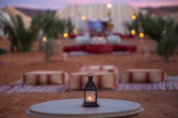 A vibrant desert camp with traditional Moroccan tents, adorned with colorful rugs and cushions. Palm trees and drums create a lively atmosphere, set against a backdrop of rolling hills and a clear blu