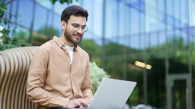 Handsome businessman works typing on a laptop while sitting on bench near a business office building. Entrepreneur chatting online, typing a message, checking email, corresponding remotely with client