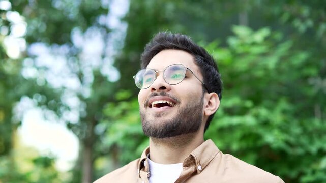 Happy joyful man relaxing with closed eyes standing in urban city park. A smiling handsome bearded male in glasses breathes deeply and enjoys being in nature. Healthy lifestyle concept. Close up