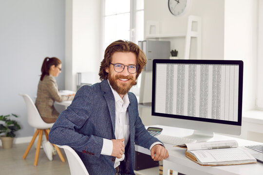 Young attractive bearded Caucasian man data science specialist smiling and looking at camera posing next to working computer with tabular data from banking industry sits at table in modern office