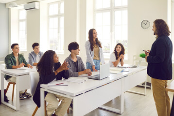 Young girl student standing at the desk in classroom and answering the male teacher with a college or high school students applauding her during a lesson. Education and learning concept.