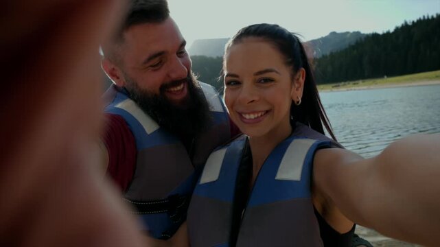 Happy couple wearing life jackets taking a selfie with a lake and mountains in the background. Smiling Couple Taking a Selfie by the Lake