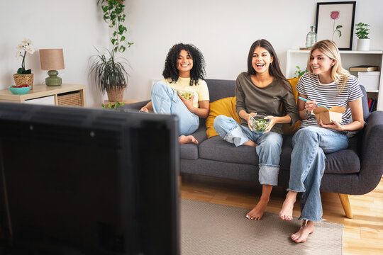 Three flatmate girl friends having fun at home watching TV movie series show while eating healthy meal 