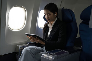 Businesswomen reading business report about finance of new startup project on tablet and listening music with bluetooth headphones while sitting near windows during flight business travel in airplane