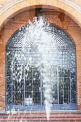 Porta com parede de tijolos, grade de ferro, chafariz esguichando água e escadaria.  
