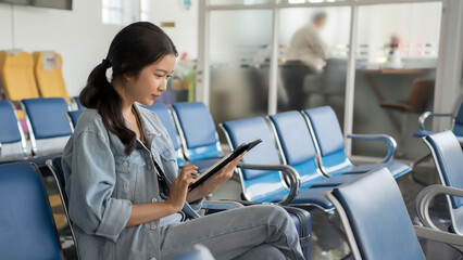 Asian traveler woman use tablet to surfing social media and typing to chat message while sitting on bench in terminal airport waiting to travelling vacation lifestyle by business travel class flight