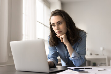 Young serious student woman or freelancer using laptop, lean at desk look at screen, read e-mail, studying or working on-line, review exam test results, consider received information, working remotely