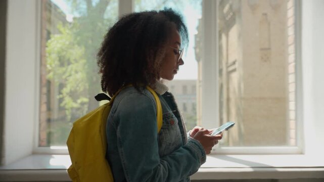 Side view African American student girl female schoolgirl woman lady walking in corridor college high school university hall walk go move going to class education study holding mobile phone smartphone