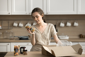 Young woman unpack carton box with ordered products, holds bottle, reads ingredients on jar with salty mix savory snacks, ordered favourite appetizers preparing for party in kitchen. Delivery services