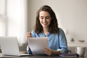 Smiling attractive student woman sit at desk with laptop, read essay, makes correction, feels...