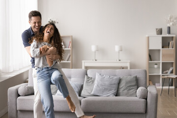 Laughing spouses hugging, dancing, having fun in living room. Playful and affectionate moment between light-hearted couple at home. Partners enjoy their romantic relationship in cozy domestic setting