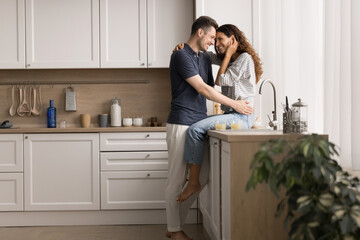 Young married couple sharing tender moment in modern kitchen. Pretty woman sitting on counter while her husband man stands close to her, gently hugging and looking into her eyes with love and warmth