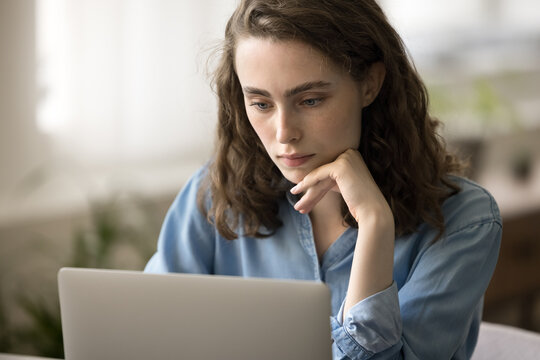 Close up serious pretty woman looks at laptop screen, ponder on answer, lead correspondence to client working remotely at home office. Student girl using internet resources studying, e-learns on-line