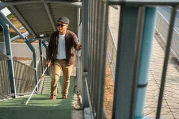 Young blind man walking in city with walking stick. Visually impaired man difficult to traveling on the road wearing sun glasses Cross the road cross the footbridge